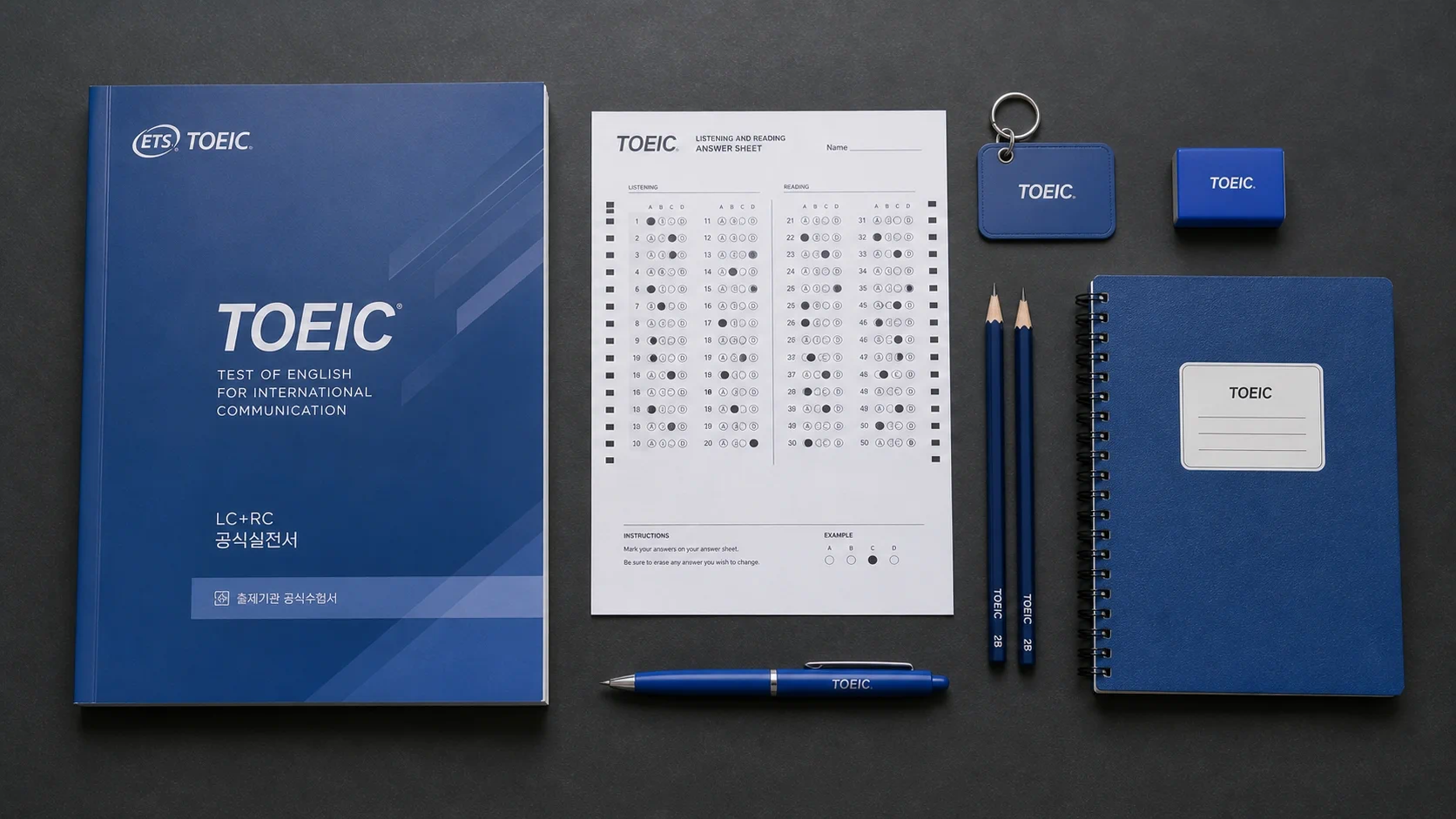 어두운 회색 책상 위에 TOEIC 교재, OMR 답안지, 파란색 키체인 카드, 연필 2자루, 지우개, TOEIC 로고가 있는 파란색 노트, TOEIC 펜이 플랫레이 구도로 배치되어 있으며, 교재 표지에 'TOEIC TEST OF ENGLISH FOR INTERNATIONAL COMMUNICATION' 텍스트가 표시됨. 미니멀하고 전문적인 시험 준비 분위기.