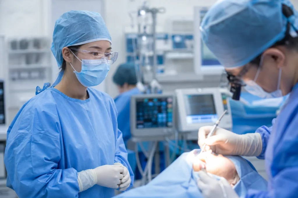 수술모로 머리를 완전히 덮고 연한 블루 또는 그린 수술복과 멸균 수술 가운을 입고 수술 마스크와 보호 안경이나 페이스 실드를 착용하고 멸균 장갑을 낀 여학생 치의학과 학생이 구강외과 수술실에서 수술 의자 옆에 서서 수술 과정을 관찰하고 있는 모습. 관찰 거리를 유지하며 손을 앞에서 모으거나 관찰 클립보드를 들고 있음. 앞쪽으로 흐릿하게 40-50대 남성 구강외과 전문의가 완전한 수술복 차림으로 실제 환자(성인, 수술 드레이프로 덮여 구강 부위만 노출)에게 수술 시술 중. 수술등이 수술 부위를 밝게 비추고 트레이에 수술 기구들이 보이며 매우 흐릿한 구강외과 수술실 배경.