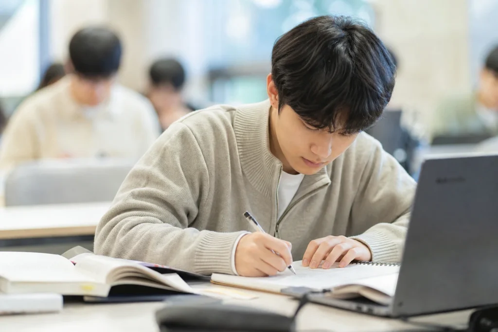 대학생이 강의실이나 학습 공간에 앉아 강렬한 집중력으로 적극적으로 상세한 노트를 작성하고 있는 집중된 사진. 소프트 베이지, 그레이, 또는 네이비 스웨터나 가디건을 입고, 매우 집중적이고 참여적인 표정으로 노트를 내려다보며 약간 앞으로 기울어져 중요한 정보를 포착하는 데 완전히 몰두하고 있음을 보여준다.